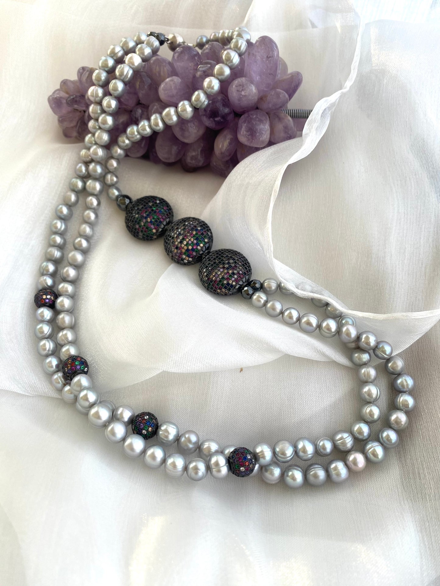 Grey Pearl Necklace with double strands of pearls arranged beside amethyst ornament on white fabric background.