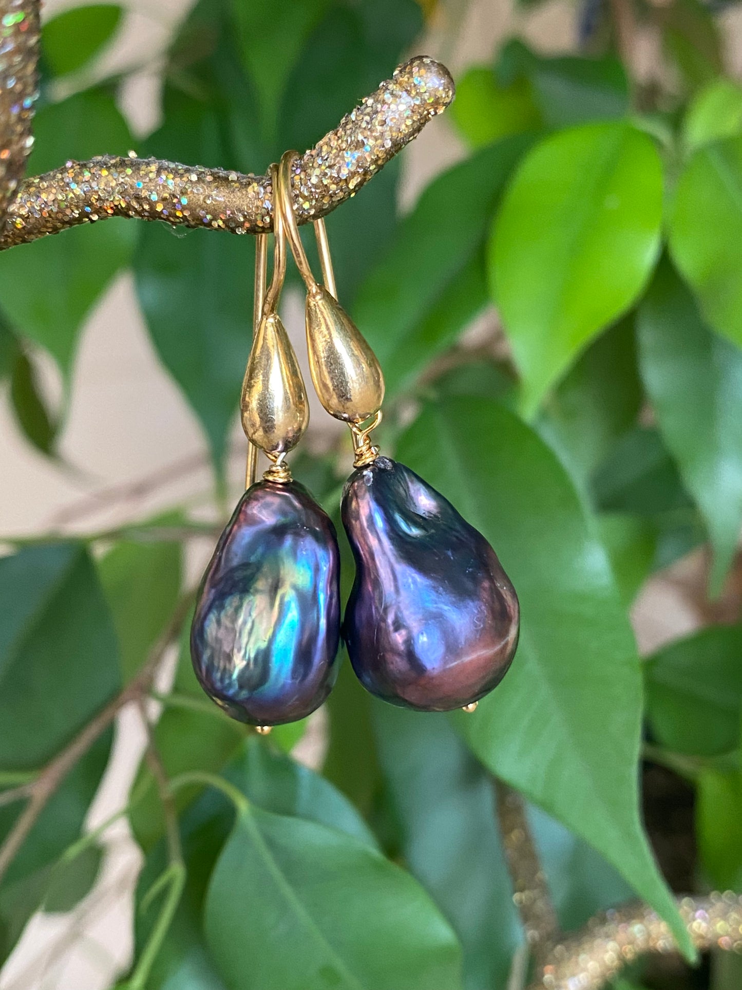 Baroque pearl drop earrings in black peacock hues held in hand against green leaf background