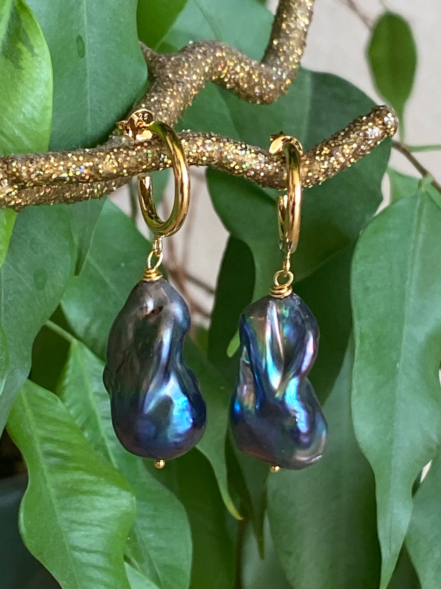 Handmade baroque pearl hoop earrings in black peacock tones shown close up on green leaf background