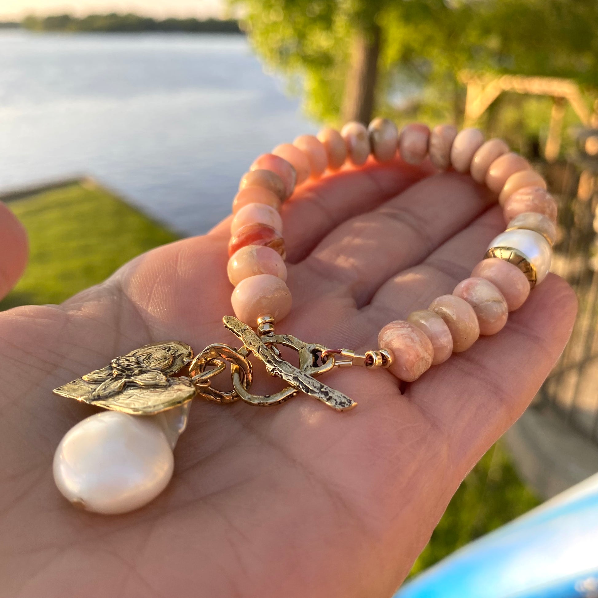 Pink opal bracelet with heart and baroque pearl charm displayed on a hand by the water.