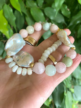 Charger l&#39;image dans la galerie, Hand holding morganite and jade necklace with baroque pearls against green natural foliage, showing gemstone color and luster.
