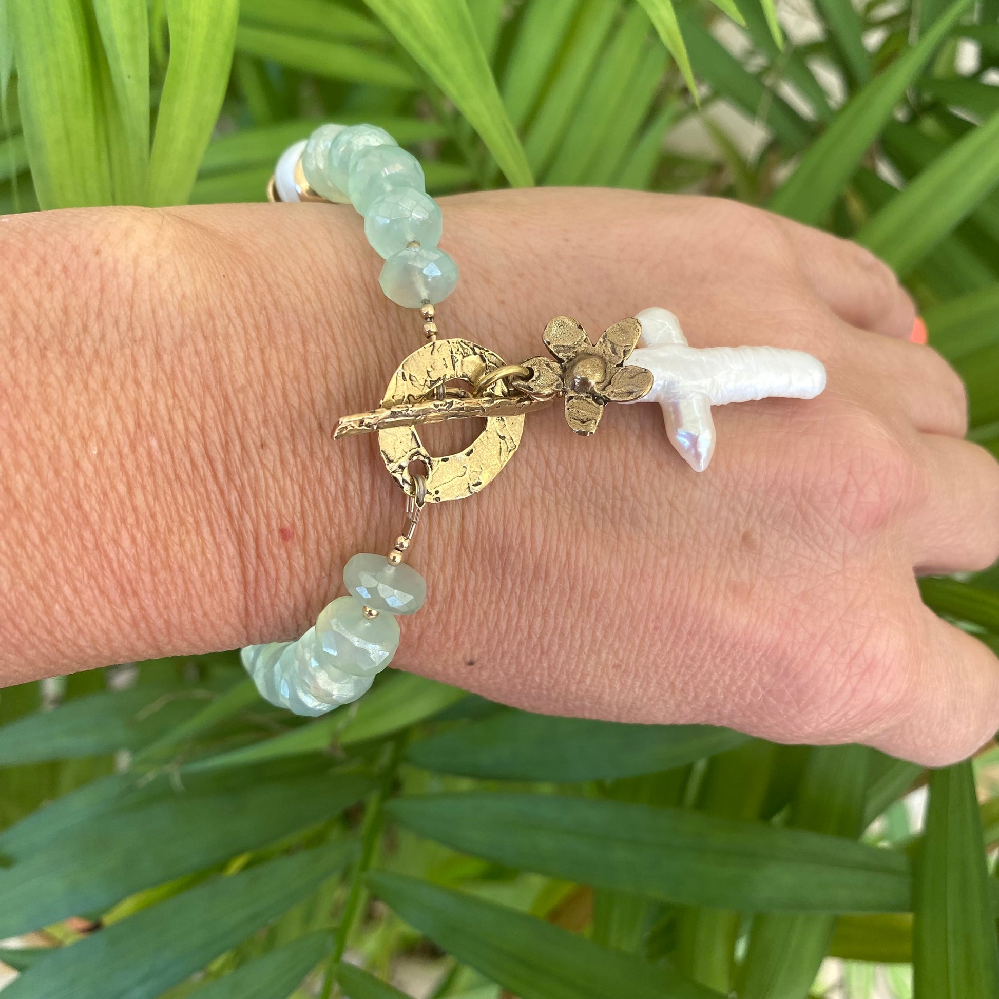 Chalcedony bracelet with cross pearl pendant and daisy charm on a hand against green foliage.