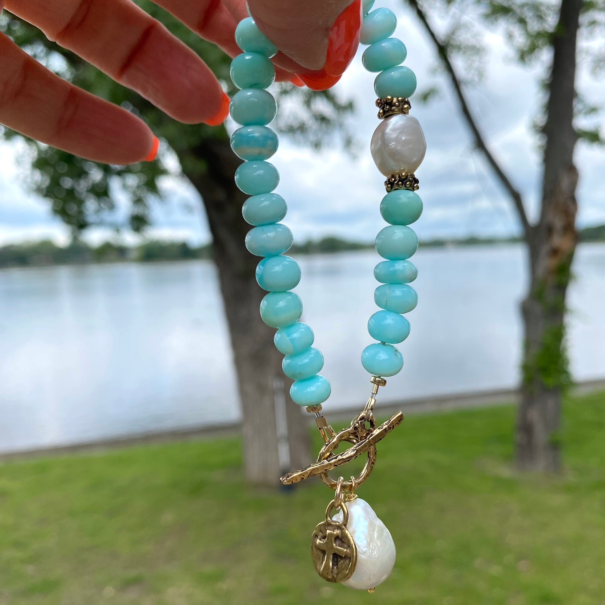 Sky blue opal bracelet with baroque pearls and cross charm, captured by a lakeside setting.