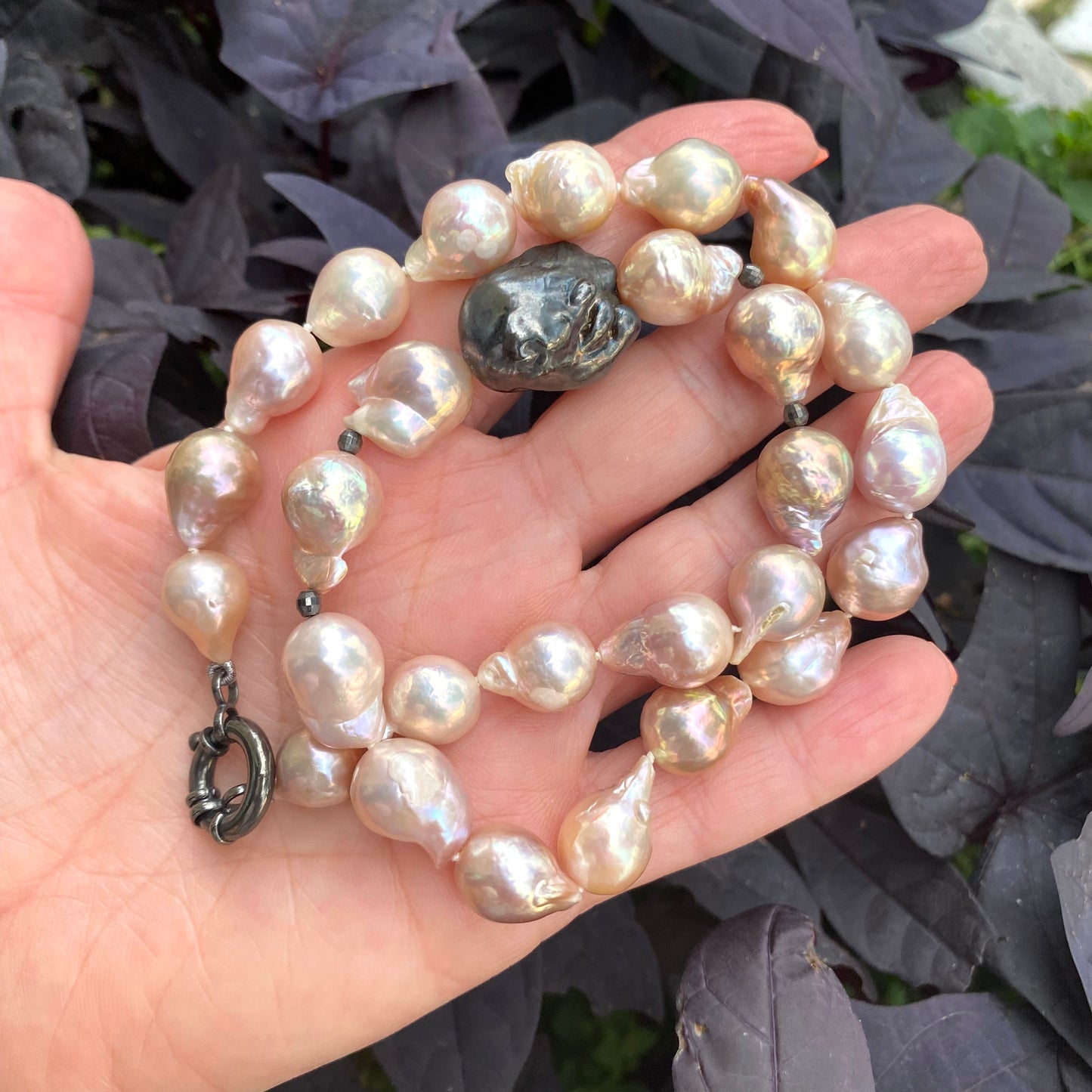 Pink baroque pearl necklace with black rhodium clasp held against purple leaves, elegant detail.