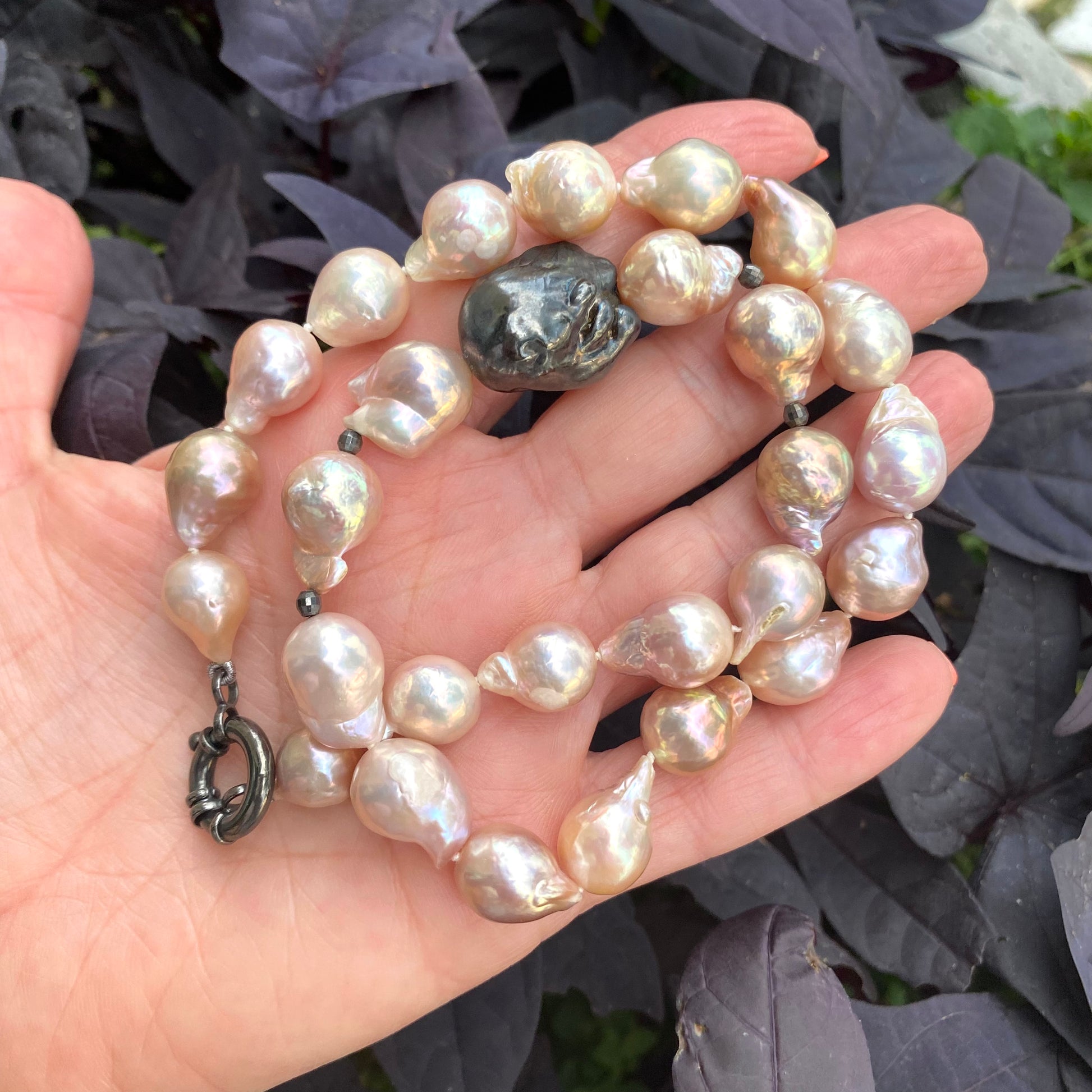 Pink baroque pearl necklace with black rhodium clasp held against purple leaves, elegant detail.