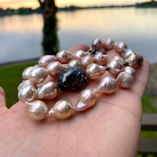 Pink baroque pearl necklace held in hand against river at sunset, shimmering metallic pearls.