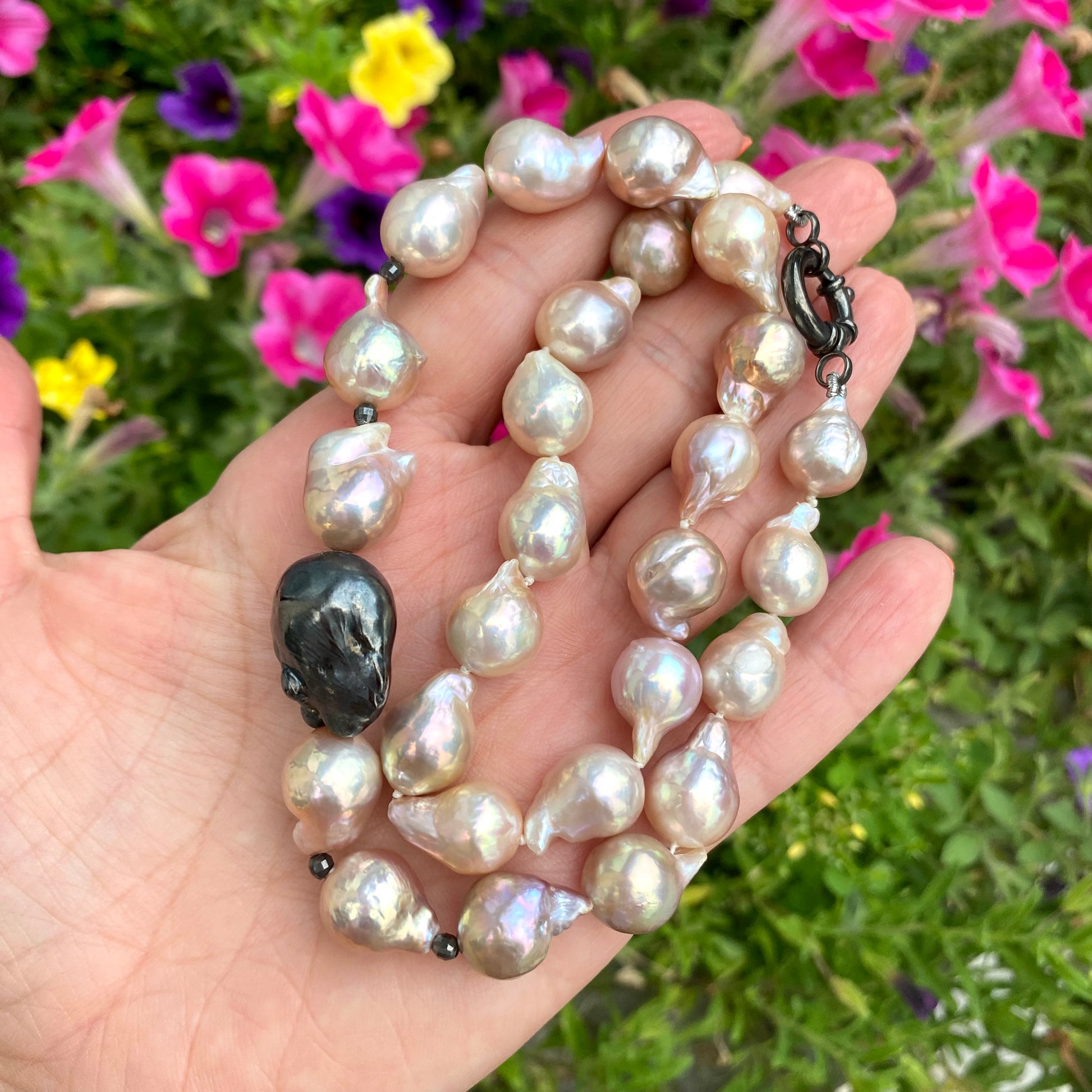 Handheld close-up of pink baroque pearl necklace against green foliage and flowers, natural light.