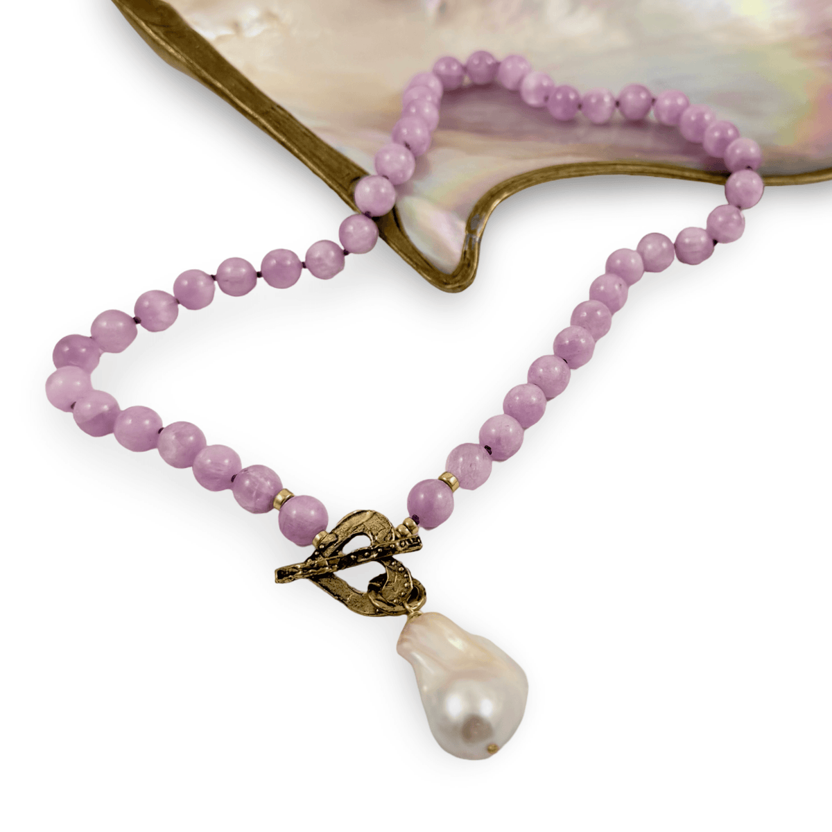 A beaded necklace with light purple/lavender round beads, a textured antique bronze clasp, and a large baroque pearl pendant, displayed on a white background with a mother-of-pearl shell prop.