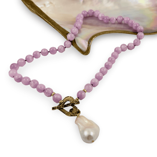 A beaded necklace with light purple/lavender round beads, a textured antique bronze clasp, and a large baroque pearl pendant, displayed on a white background with a mother-of-pearl shell prop.