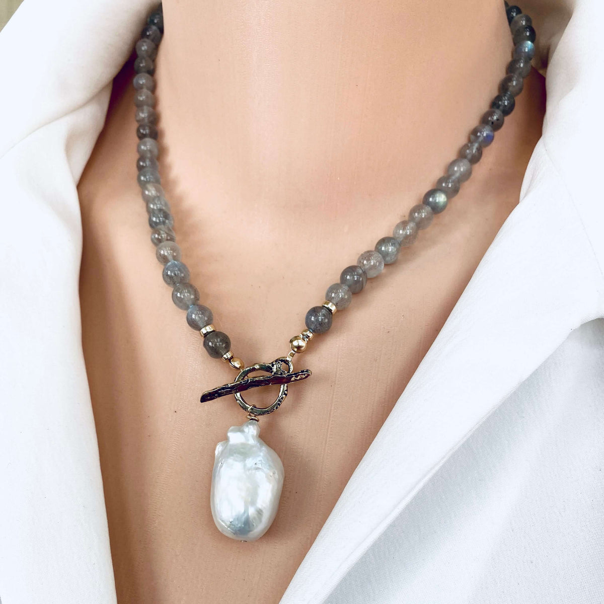 A close-up of a beaded necklace worn on a model's neck, featuring gray stone beads, small gold-toned spacer beads, a dark toggle clasp, and a large white baroque pearl pendant.