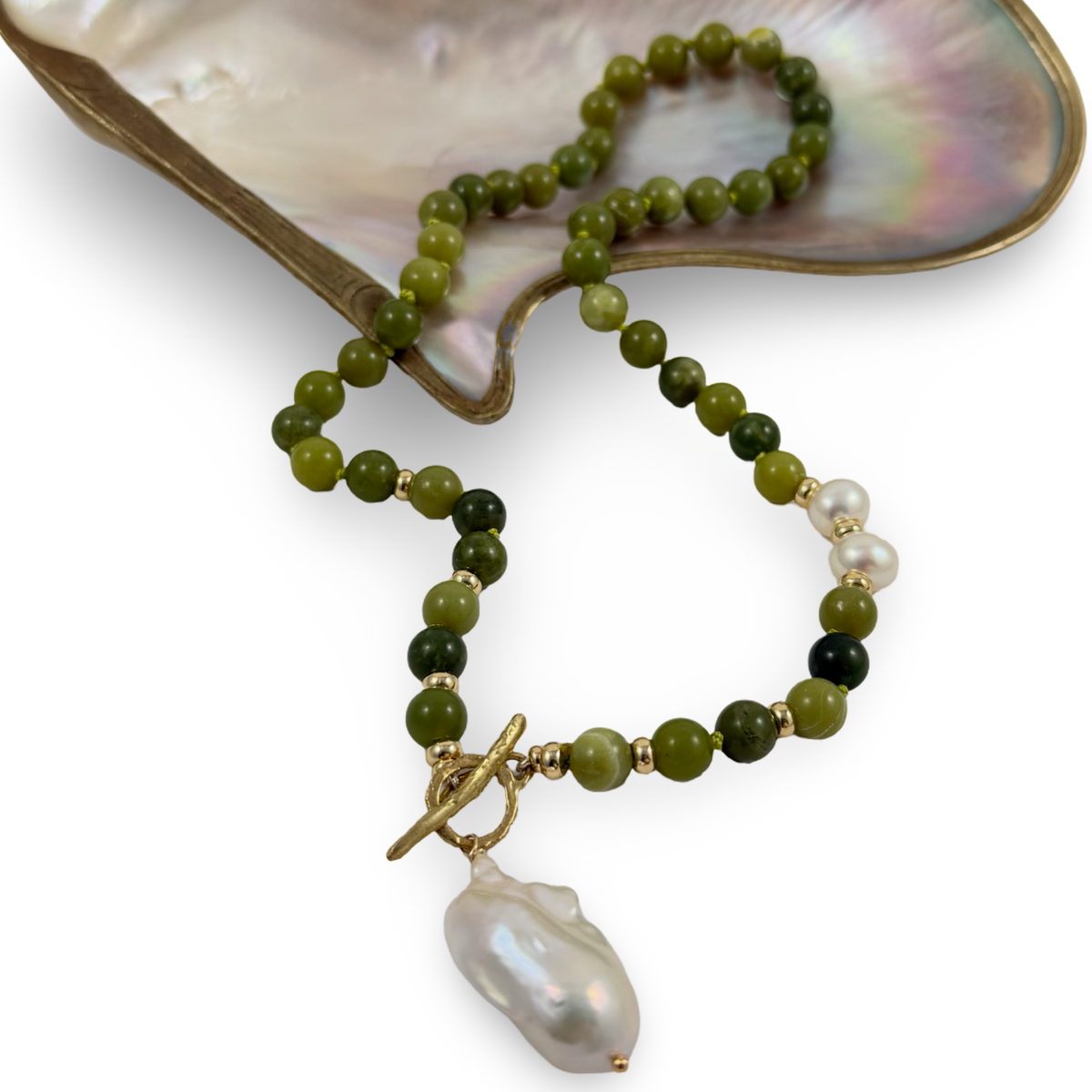 A beaded necklace with olive green and dark green round beads, accented with small gold spacer beads, a white pearl, and a large baroque white pearl pendant, displayed on a mother-of-pearl shell against a white background.