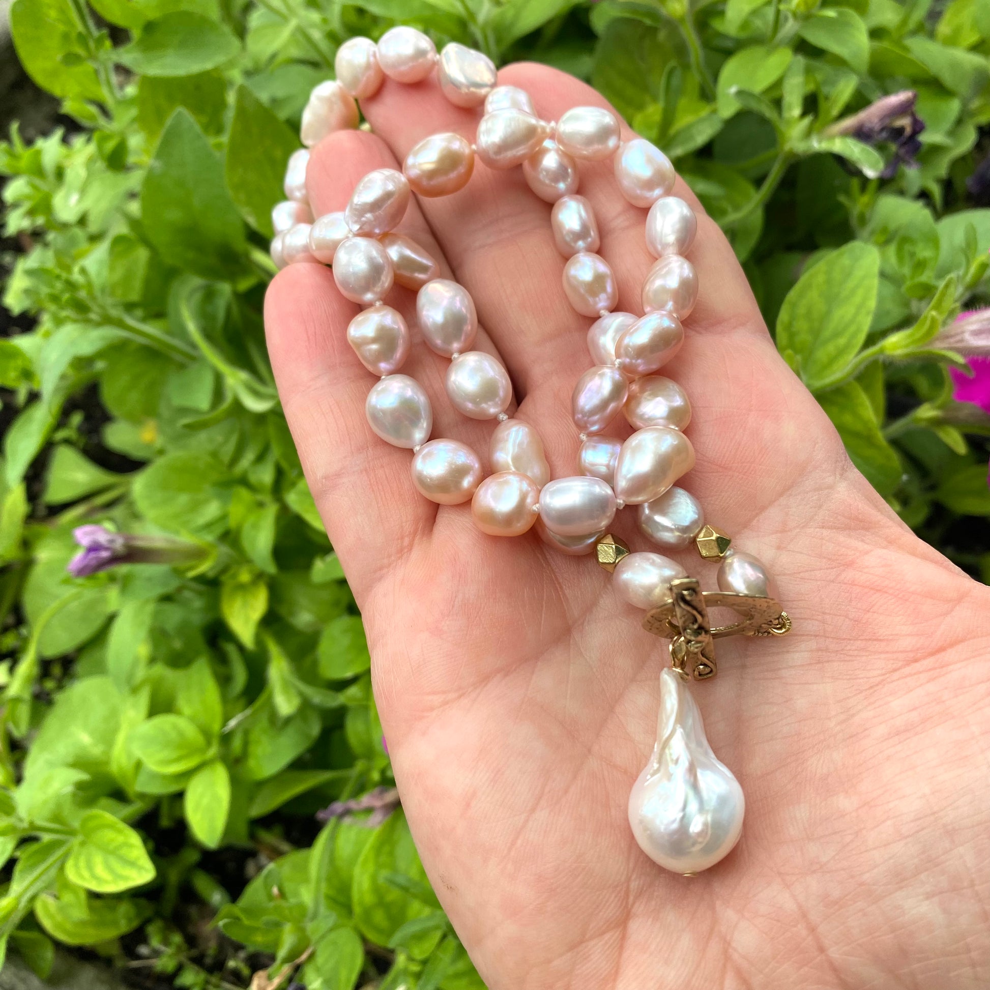 Pink pearl necklace with baroque pendant held in hand against green foliage