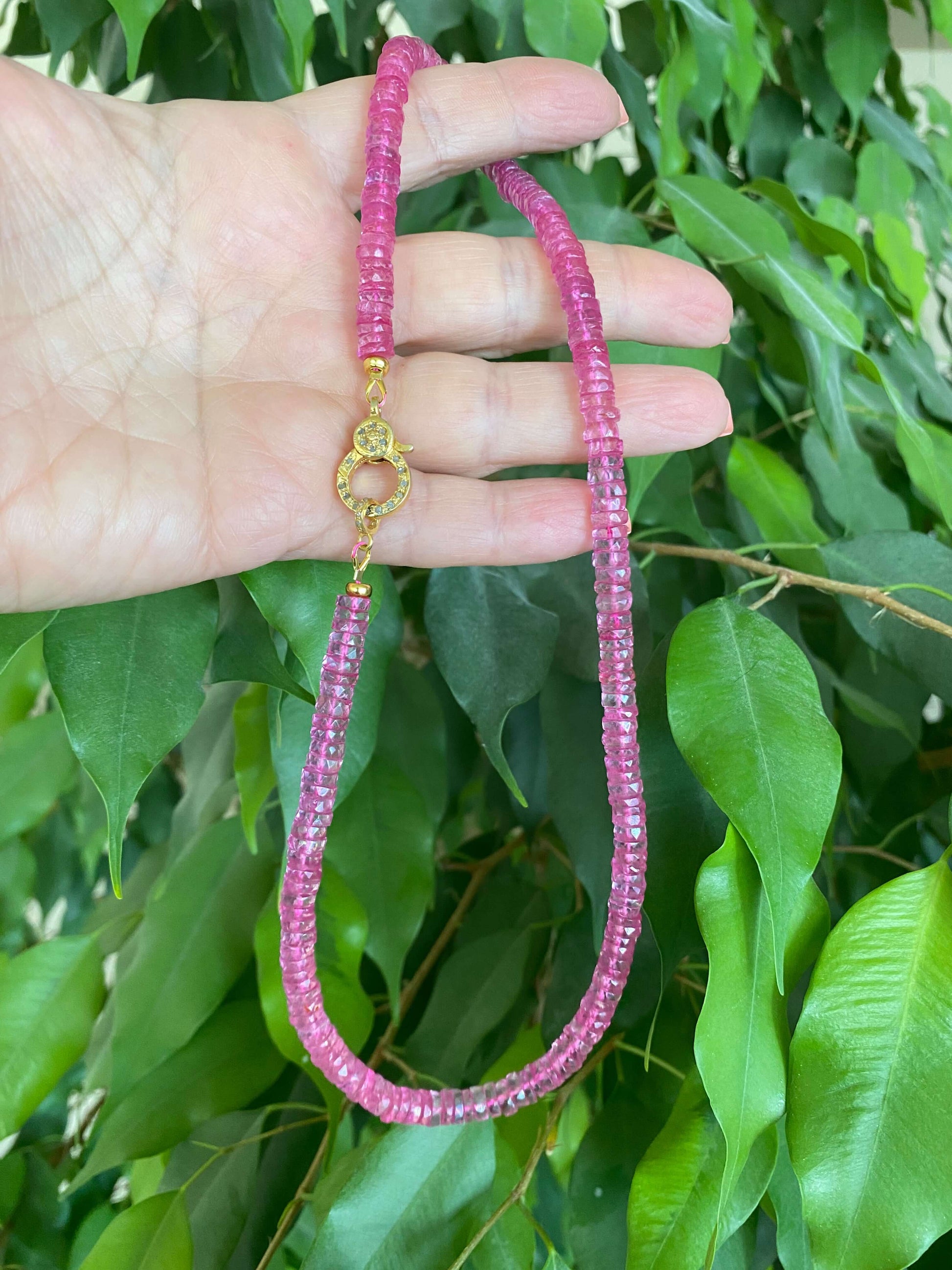 Pink topaz beaded necklace with gold vermeil pavé clasp displayed on a hand among green leaves.