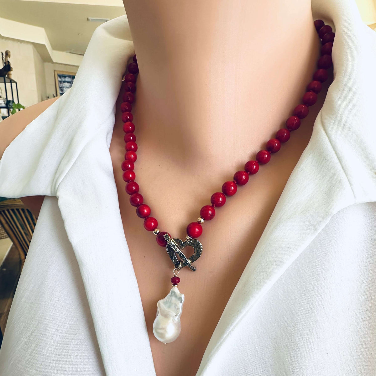A close-up of a red beaded necklace with a silver-toned heart-shaped clasp and a white baroque pearl pendant, worn on a mannequin or model with a white collared shirt.