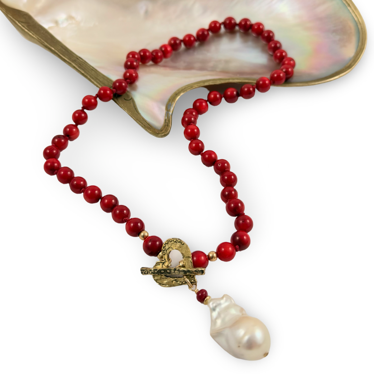 A beaded necklace made of glossy red beads with a gold-tone toggle clasp and a large white baroque pearl pendant, displayed on a mother-of-pearl shell against a white background.