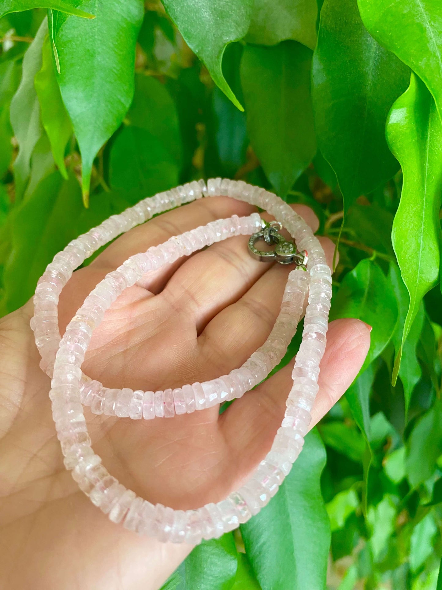 Hand holding a rose quartz beaded necklace with oxidized silver pavé clasp among lush green leaves.