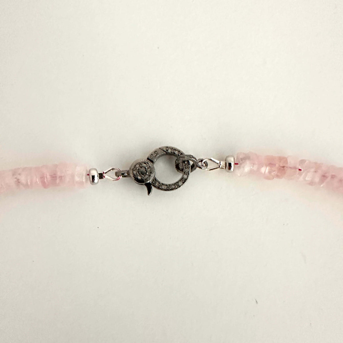 A close-up of a beaded bracelet featuring light pink faceted beads and a central dark gray textured toggle clasp on a plain off-white background.
