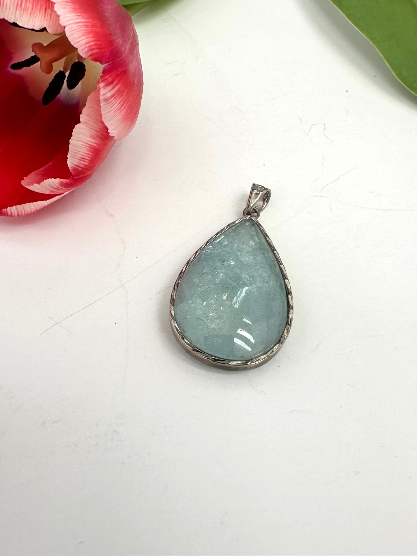Teardrop-shaped pendant with a light blue stone on a white surface next to a pink tulip.