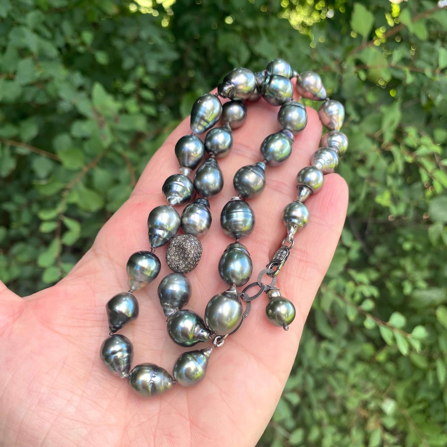 Tahitian baroque pearl necklace with champagne diamonds, featuring unique pearls and diamond accents, hand-held near greenery.