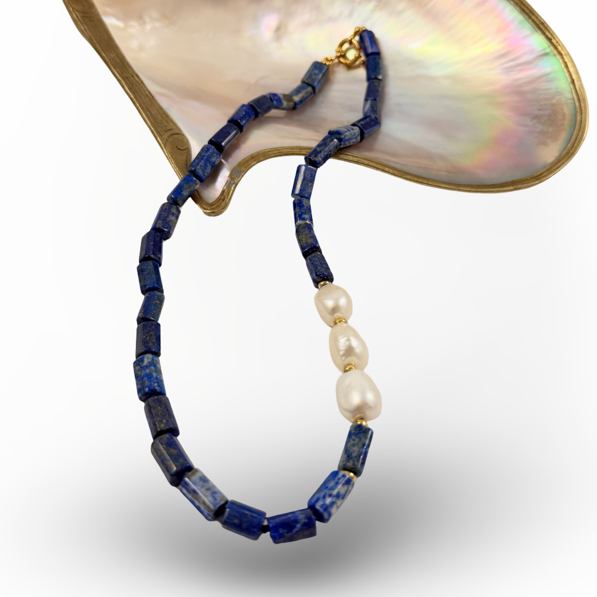 A beaded necklace with dark blue lapis lazuli-style rectangular beads, three white baroque pearls, and a gold clasp, displayed against a white background with a mother-of-pearl shell prop.