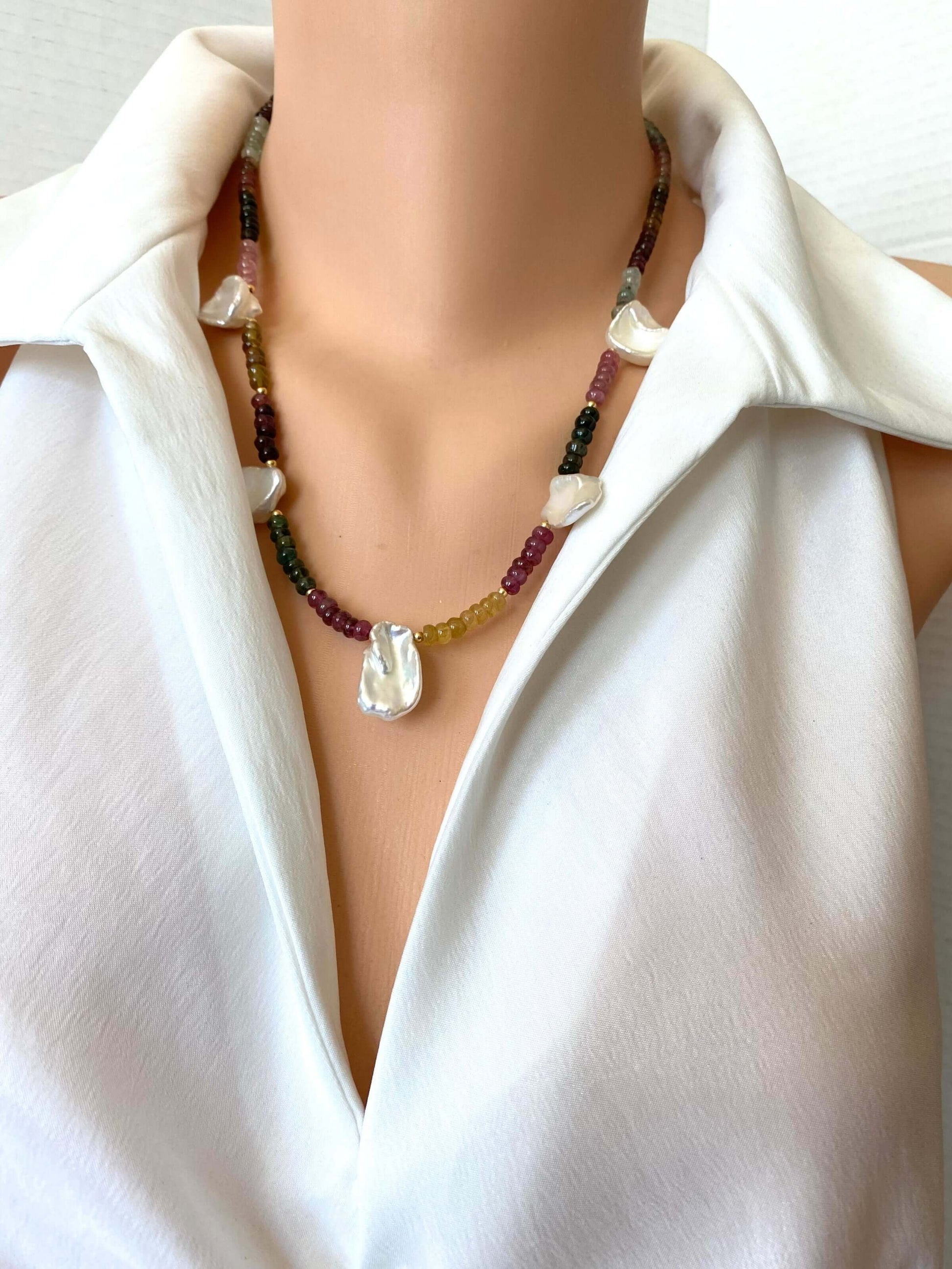 A close-up of a person wearing a multi-strand beaded necklace with a large white teardrop pendant, styled with a white collared shirt.