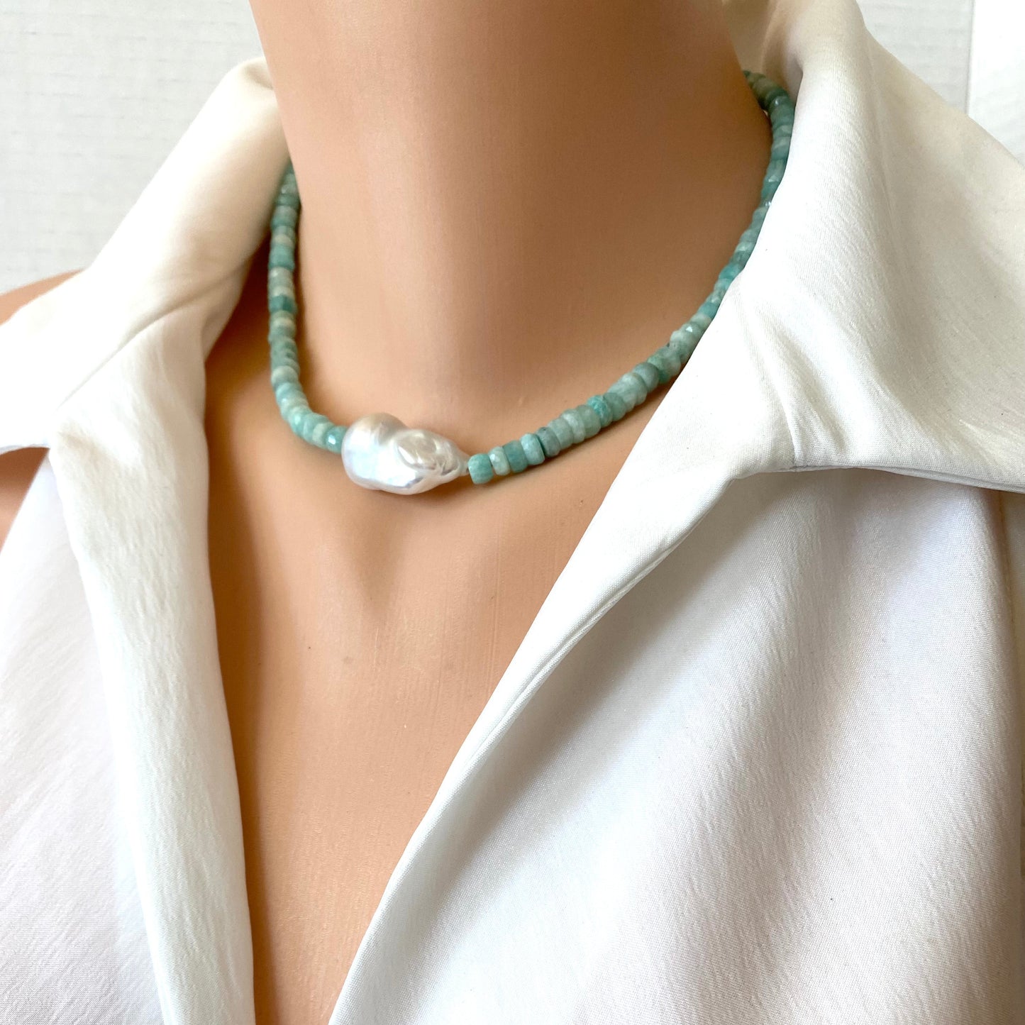 A close-up of a model wearing a light blue-green beaded necklace with a large white baroque pearl pendant, styled with a white collared shirt.