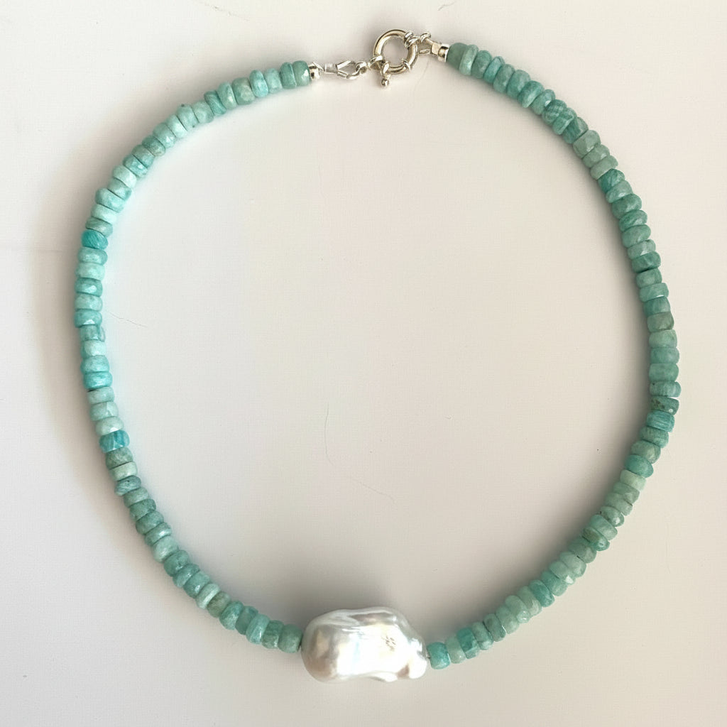 A beaded necklace featuring small, faceted turquoise-blue beads and a large, irregular white pearl pendant, displayed on a light off-white background.