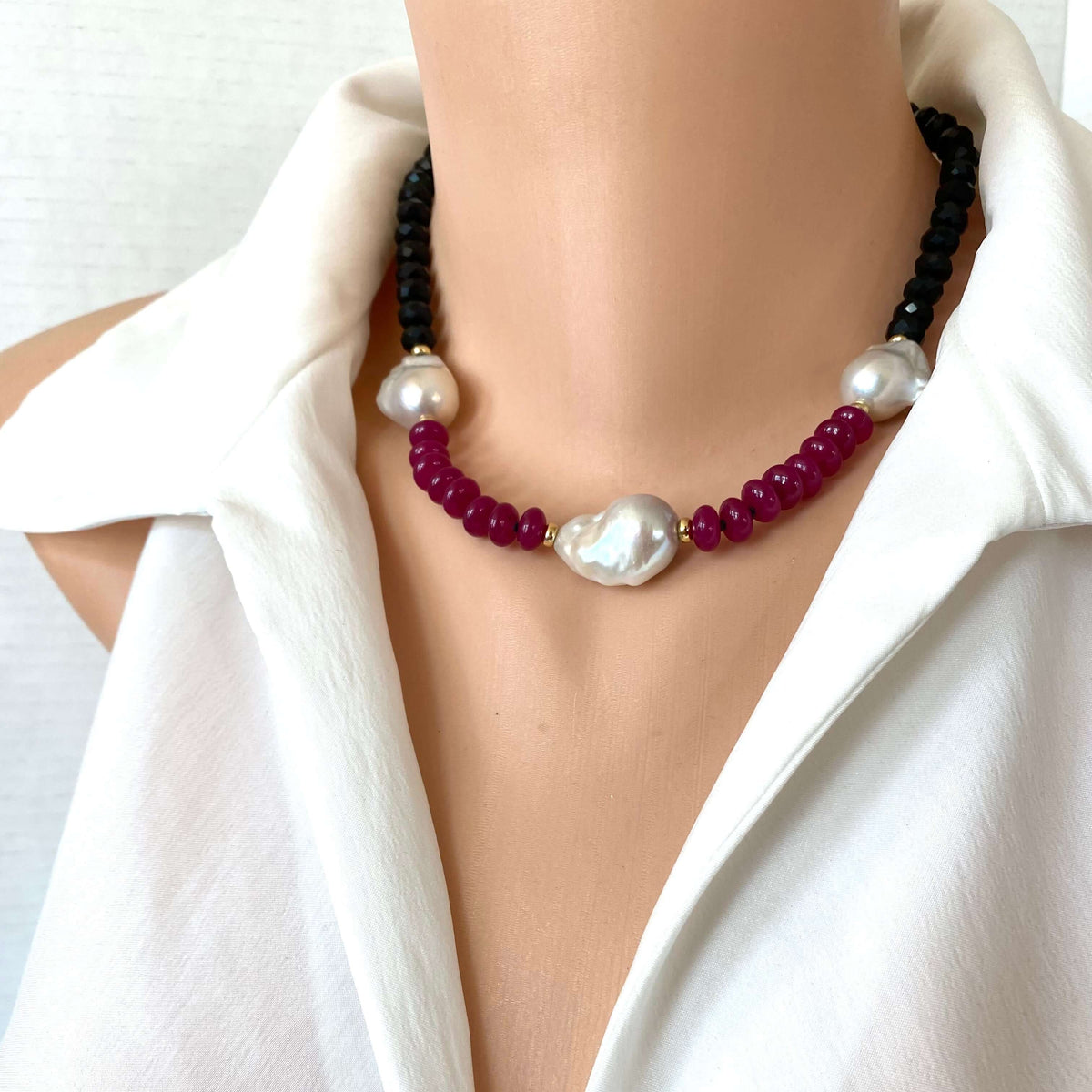 A close-up of a beaded necklace displayed on a mannequin torso wearing a white blouse, dark black beads with deep red beads, accented by white pearls.