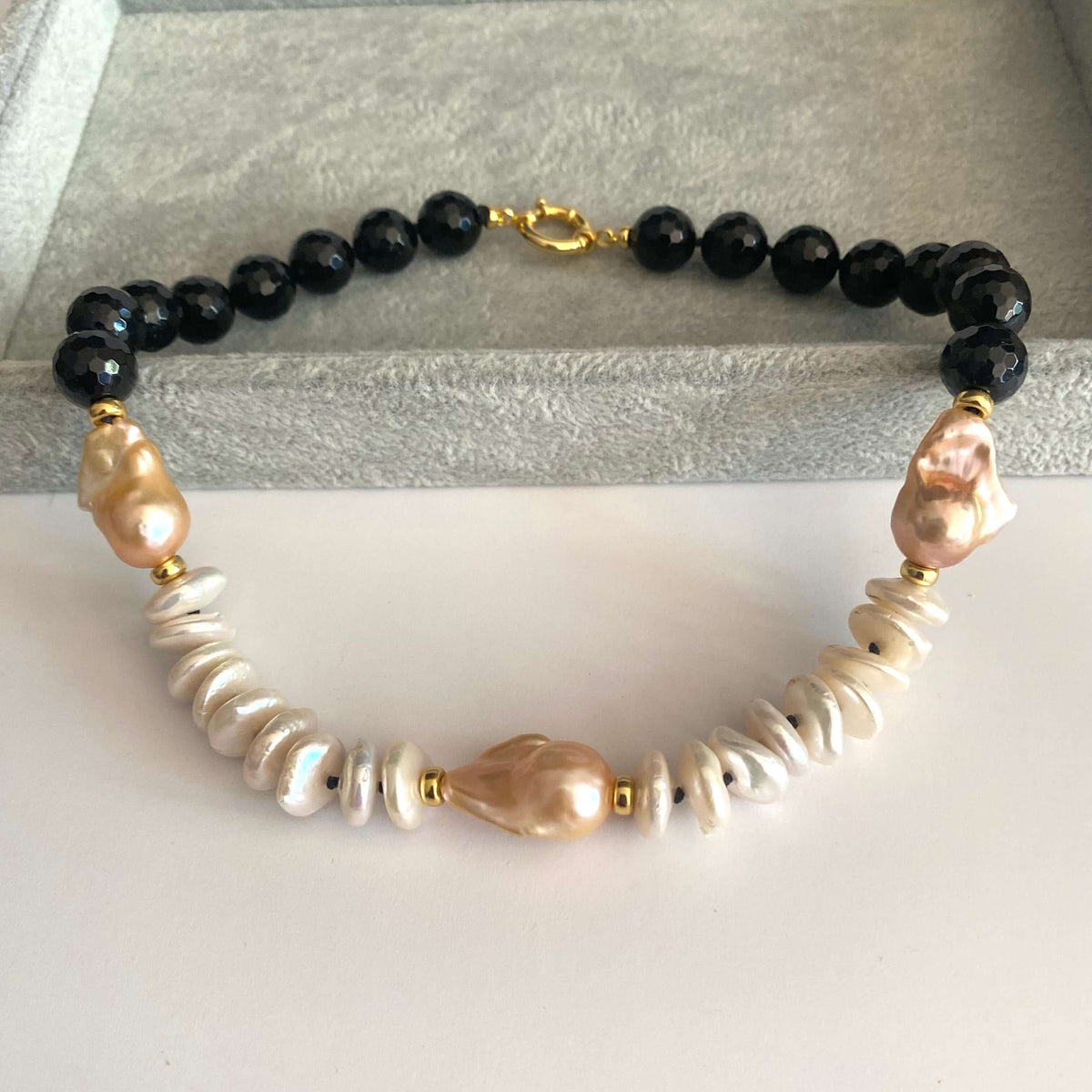 A beaded necklace featuring black faceted beads, white baroque pearls, and pale pink baroque pearls, separated by small gold spacer beads, displayed on a gray velvet jewelry box.