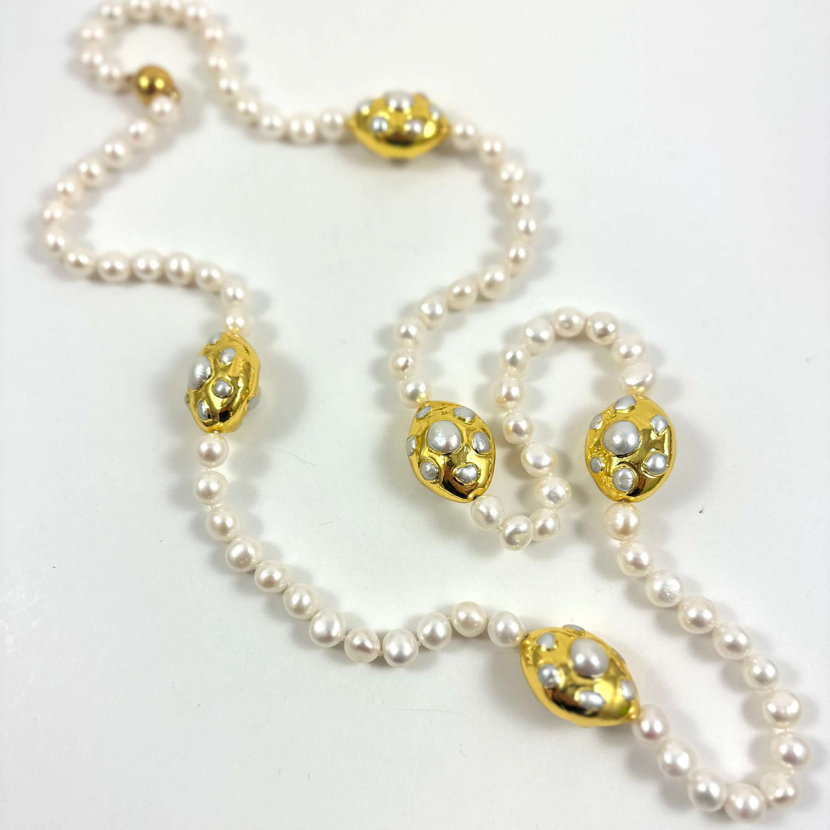 A vintage-style pearl necklace with five ornate gold-toned oval medallions, each decorated with small white pearls, laid out on a plain white surface.