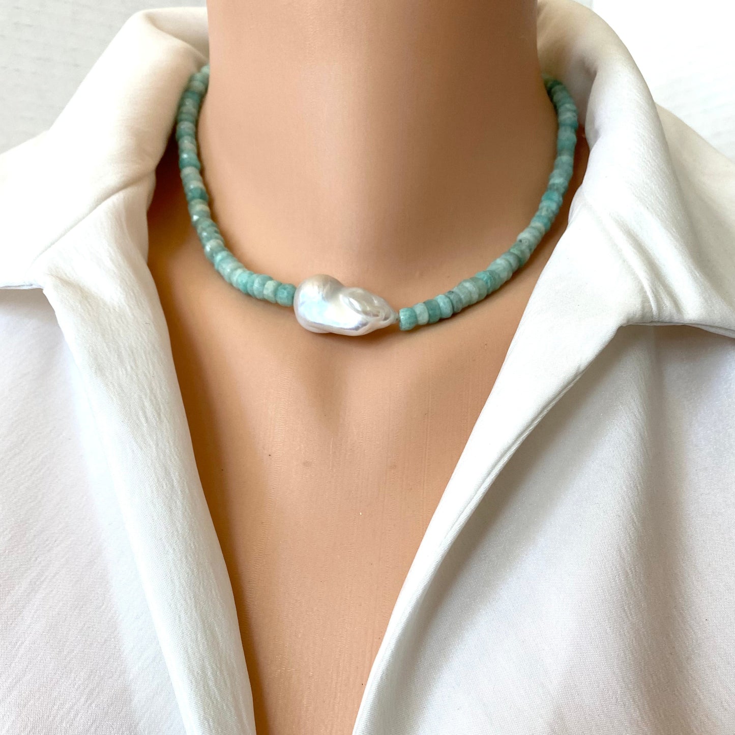 A close-up of a beaded necklace with light blue-green round beads and a large irregular white pearl pendant, worn on a mannequin or model torso in a white collared blouse.