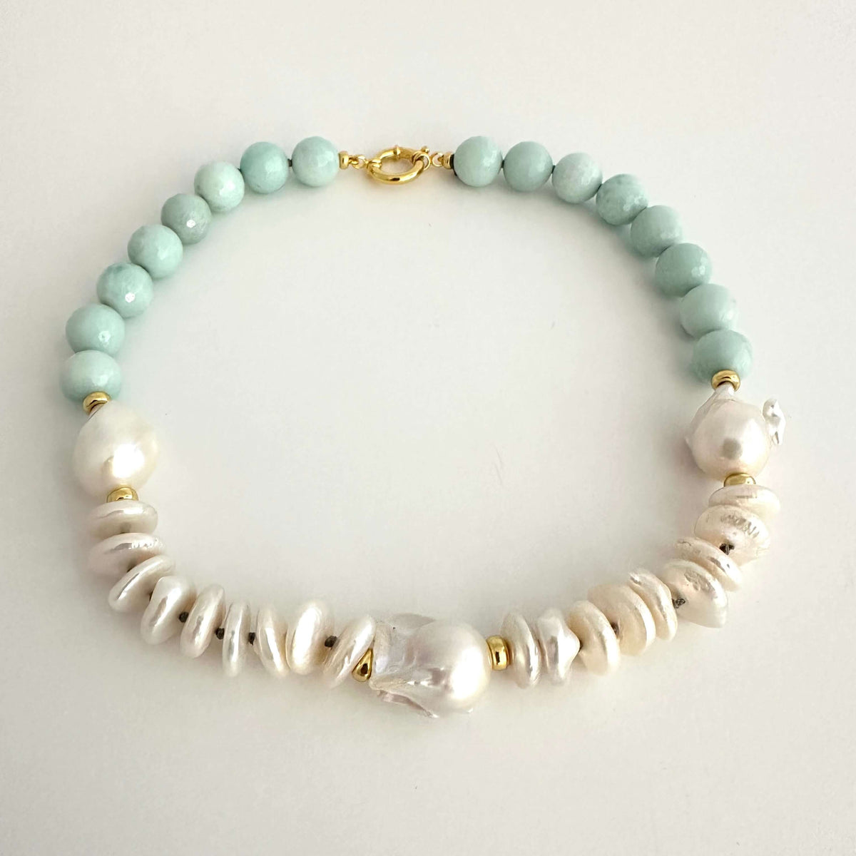 A beaded necklace featuring light mint green round beads, white baroque pearls, and textured white disc beads, accented with small gold spacer rings and a gold clasp, displayed on a light gray background.