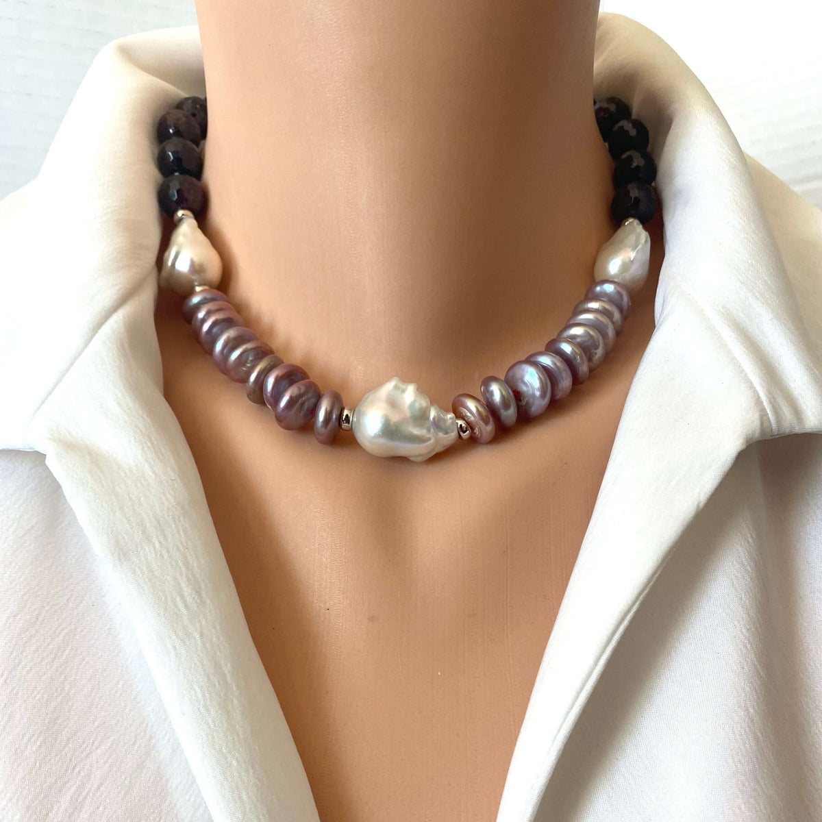 A close-up of a double-strand pearl necklace displayed on a mannequin torso wearing a white collared shirt. The necklace features dark brown beads on one side, iridescent baroque pearls on the other, and a central large white baroque pearl.