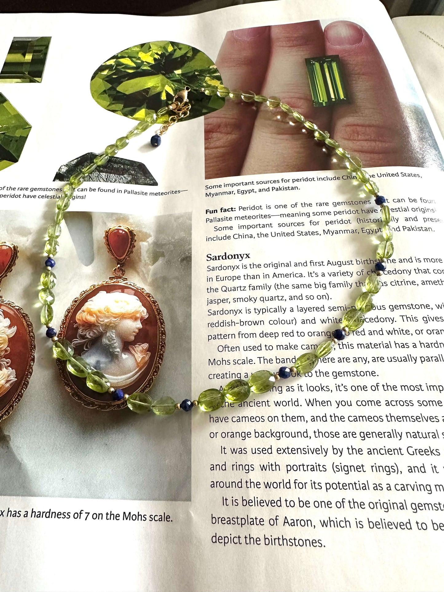 A handmade pendant featuring a green gemstone and a copper-toned textured band with red and green beads, displayed on an open book with text visible.