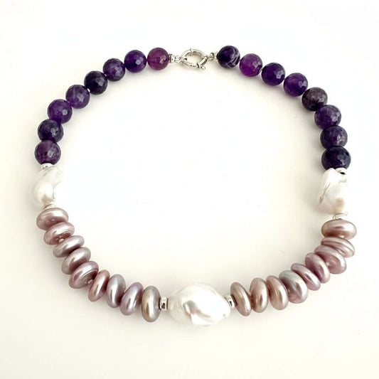 A beaded necklace featuring dark purple amethyst beads on one side, large white baroque pearls on the other, and iridescent pinkish-brown baroque pearls in the center, displayed on a plain off-white background.