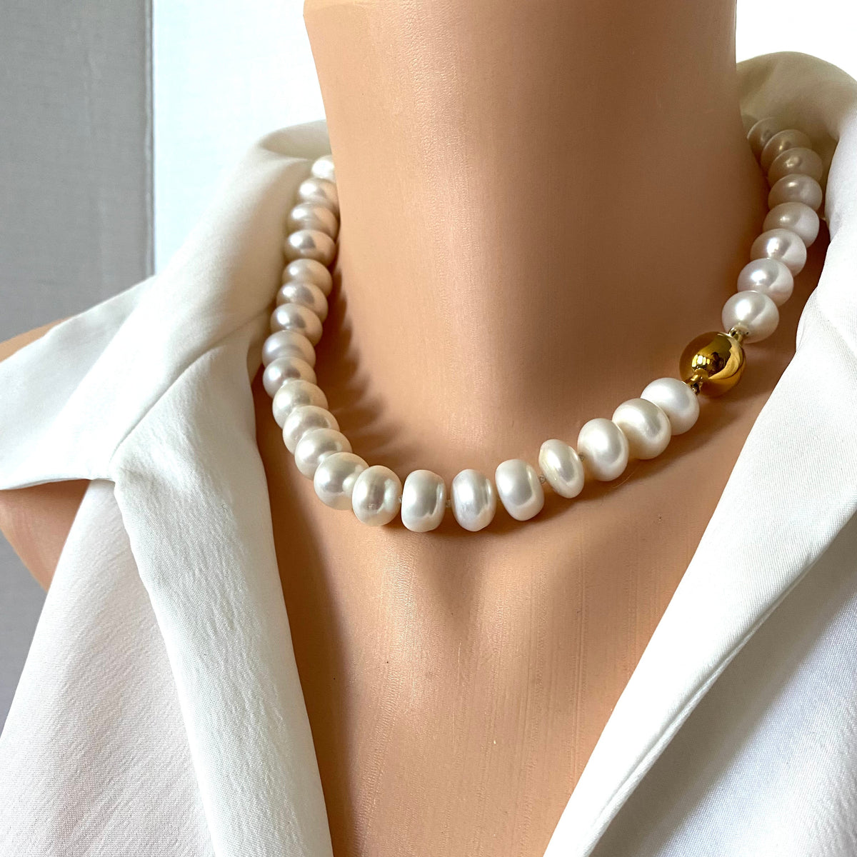 A close-up of a pearl necklace with a single gold bead accent, displayed on a mannequin torso wearing a white blouse.