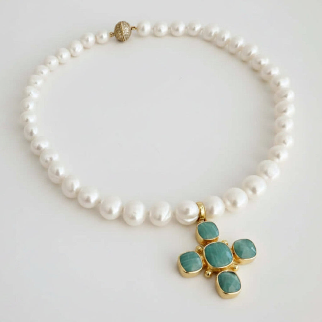 A pearl beaded necklace with a gold cross pendant featuring green cabochon stones, displayed on a light gray background.