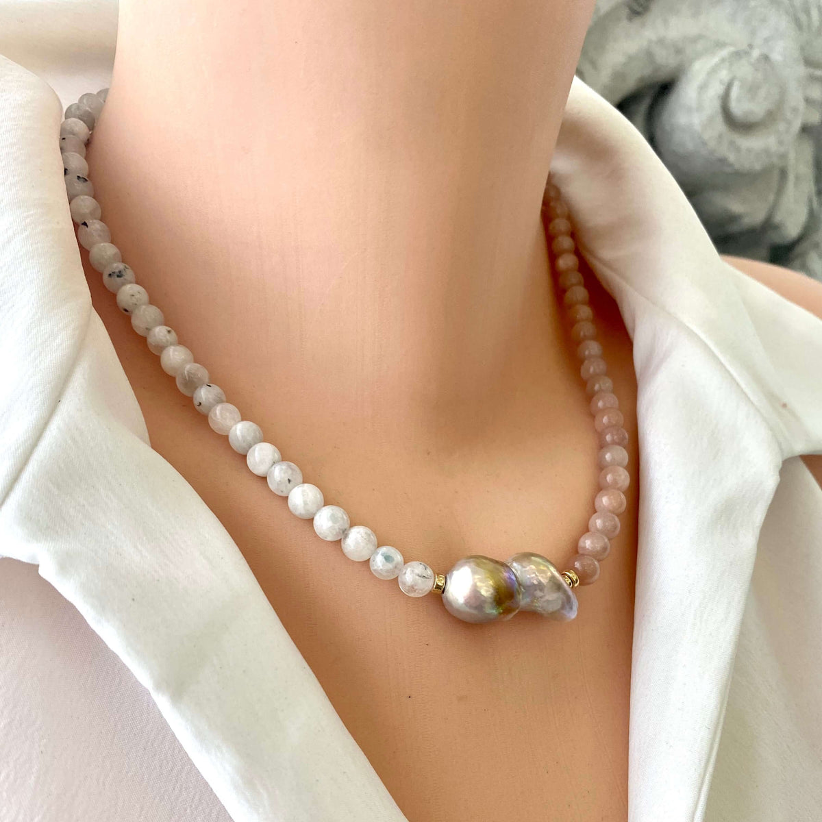 A close-up of a beaded necklace worn on a model's neck. The necklace features a strand of white marbled beads and two baroque-style pearls in iridescent gold, silver, and green tones, accented by a small gold connector.