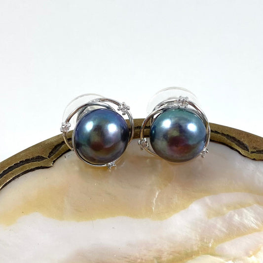 A pair of silver stud earrings featuring round, iridescent dark blue-gray pearls, displayed on a polished shell surface against a white background.
