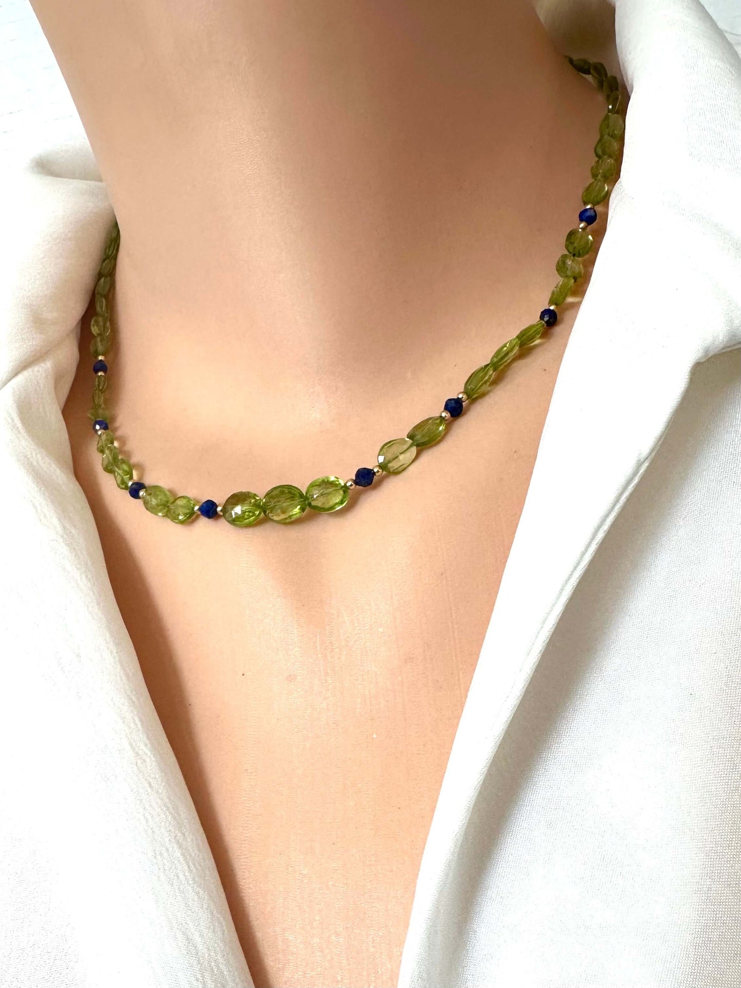 A close-up of a beaded necklace worn on a mannequin or model neck, featuring olive green faceted beads with small dark blue accent beads, styled with a white collared shirt.