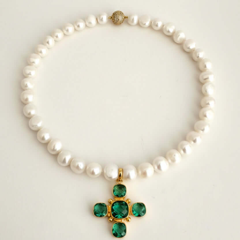 A white pearl necklace with a gold cross pendant featuring green gemstones, displayed on a light cream background.