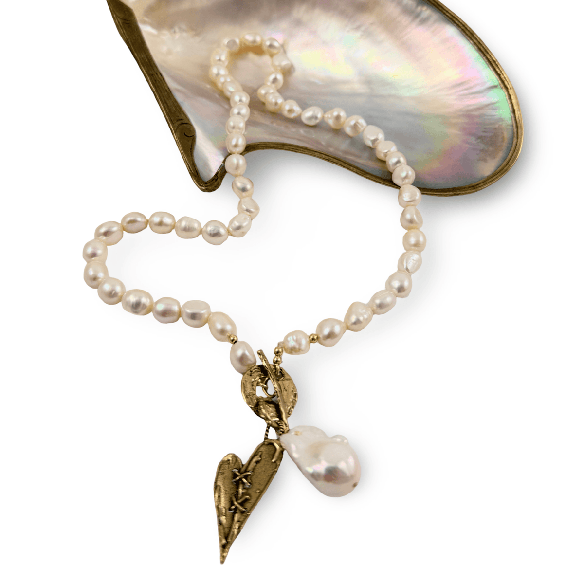 A pearl and gold necklace featuring a heart-shaped pendant with a star motif and a baroque pearl, displayed on a polished iridescent mother-of-pearl shell against a white background.