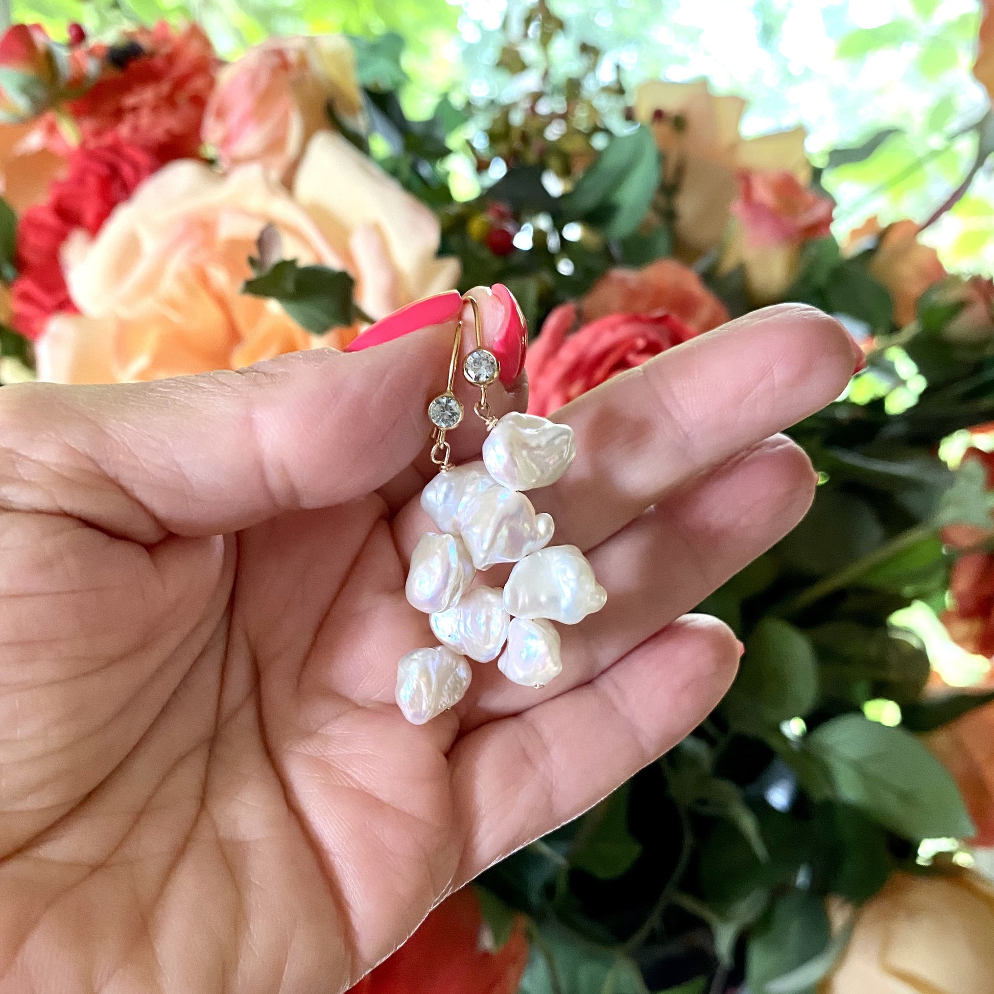 Pearl drop earrings featuring keshi pearls and blue CZ, elegantly showcased in a hand against floral backdrop.
