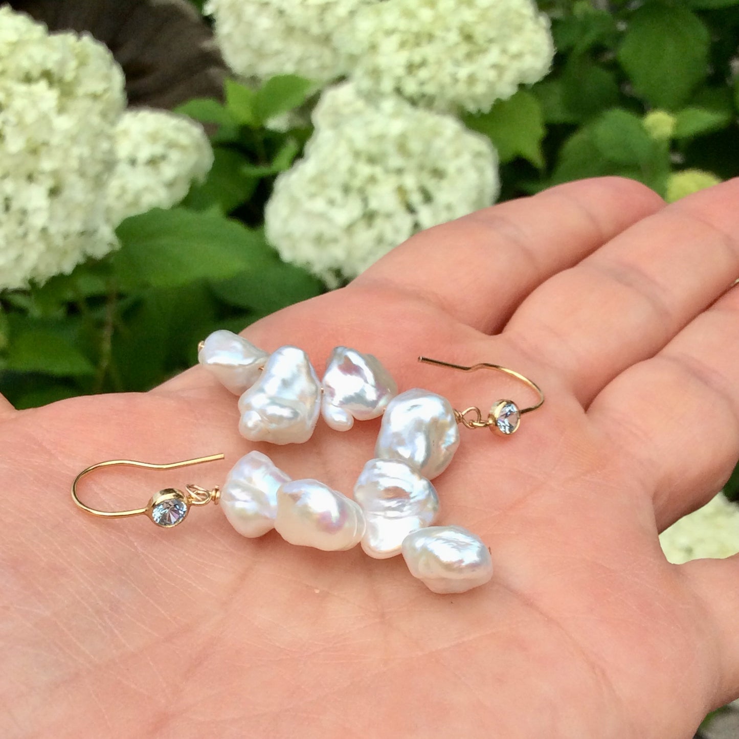 Pearl drop earrings featuring keshi pearls and blue CZ, handmade in Canada, displayed on a hand among flowers.