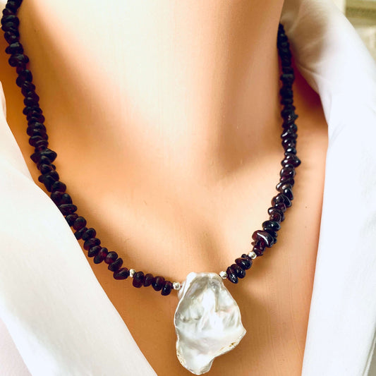 A close-up of a necklace displayed on a mannequin bust wearing a white top. The necklace features a strand of dark, red beads with a large, irregular silver pearlpendant.