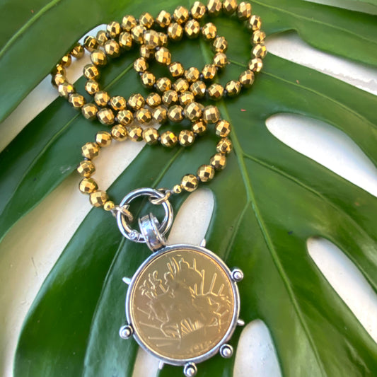 Mexican Peso Coin Pendant Necklace on a gold-coated hematite bead chain, resting on a green leaf.