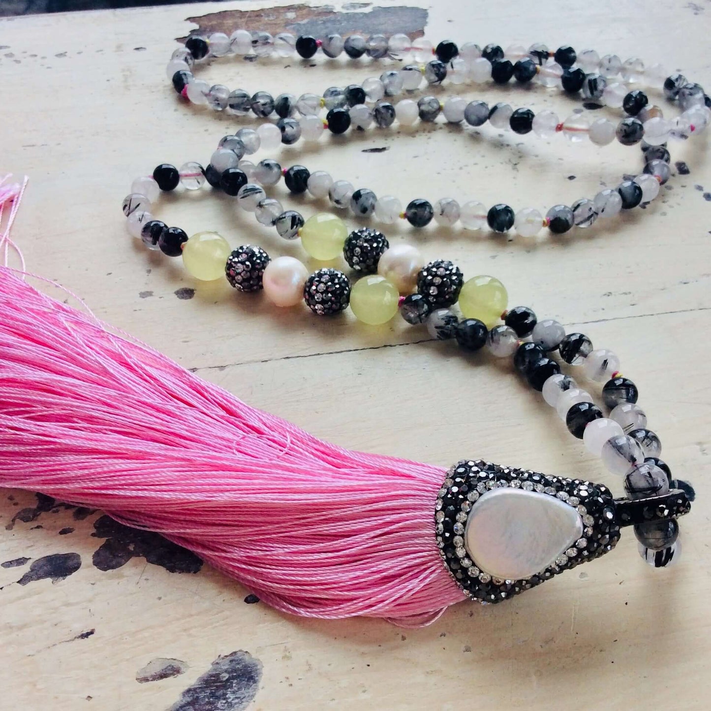 Rutilated quartz necklace with pink tassel on white wooden table, close-up view.